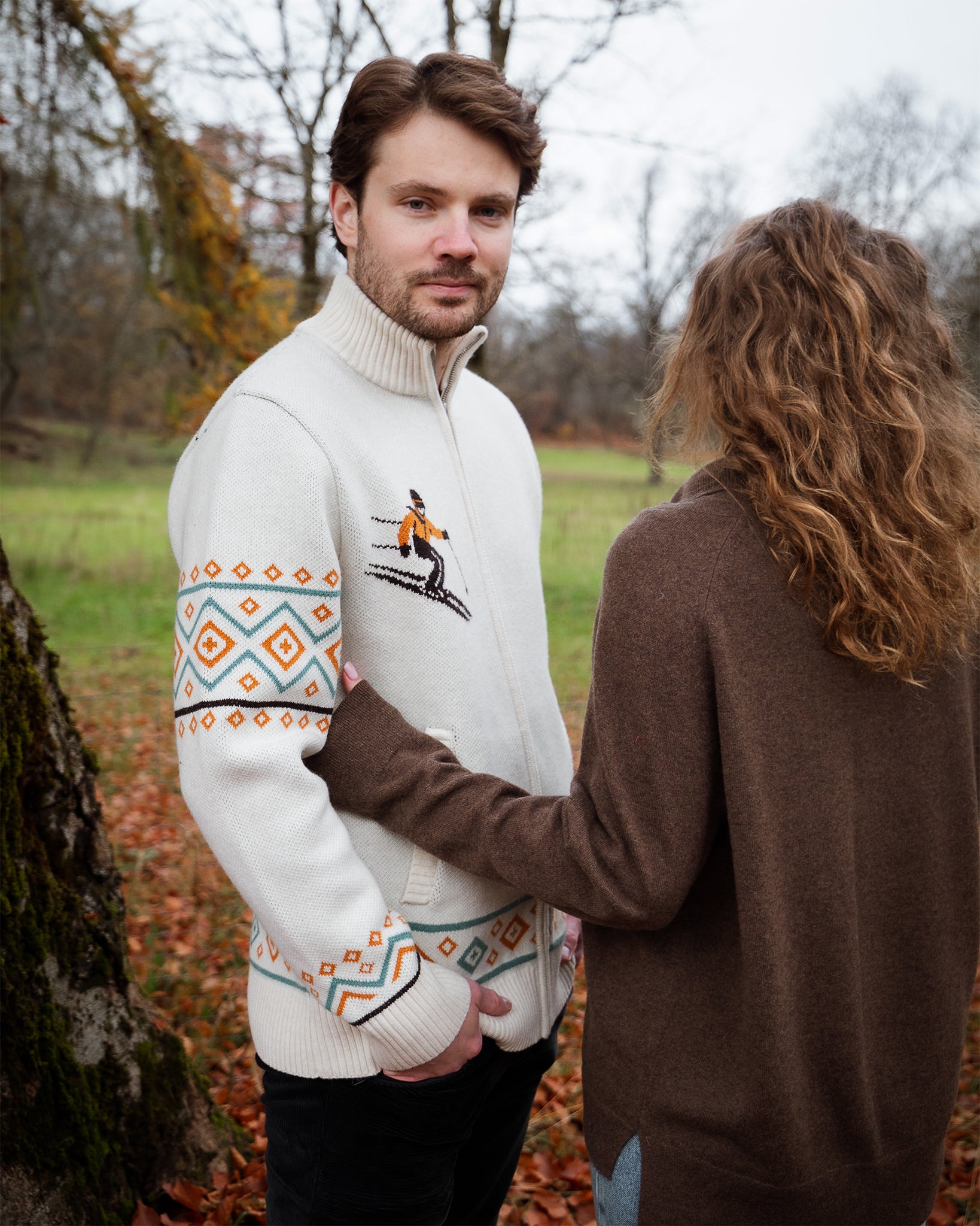 Man wearing a white sweater with colorful patterns and a logo, standing outdoors in a natural setting. A woman is standing next to him.