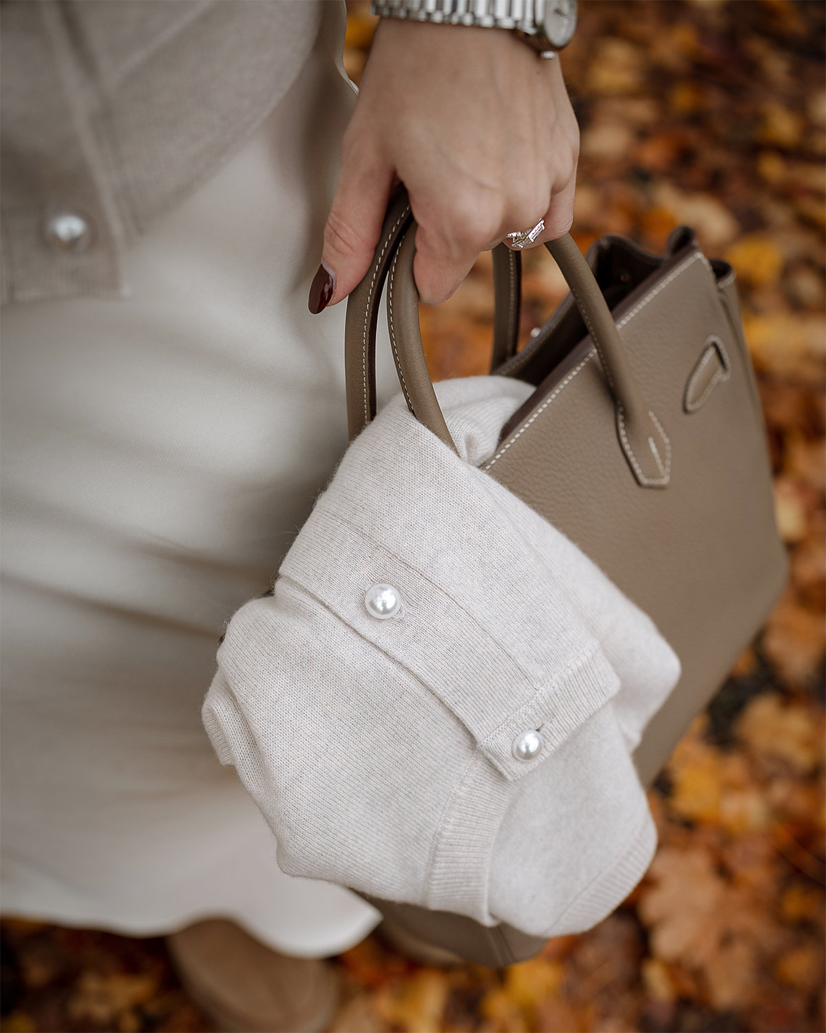 Person holding a beige handbag with a textured white pouch attached, against a blurred autumn leaf background.