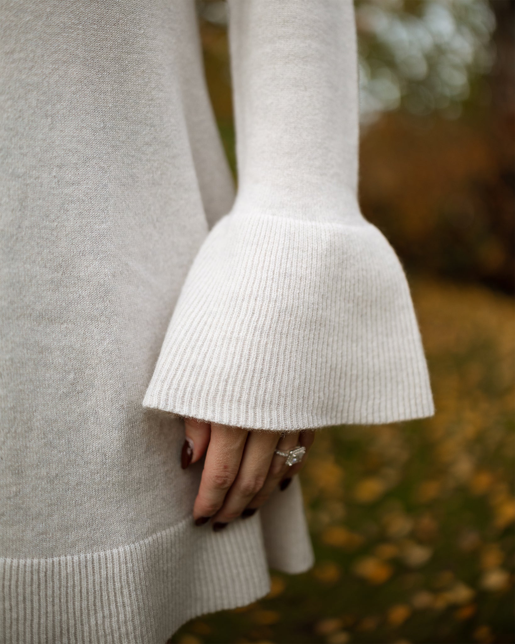 Close-up of a person wearing a light gray sweater with bell sleeves against a blurred natural background.
