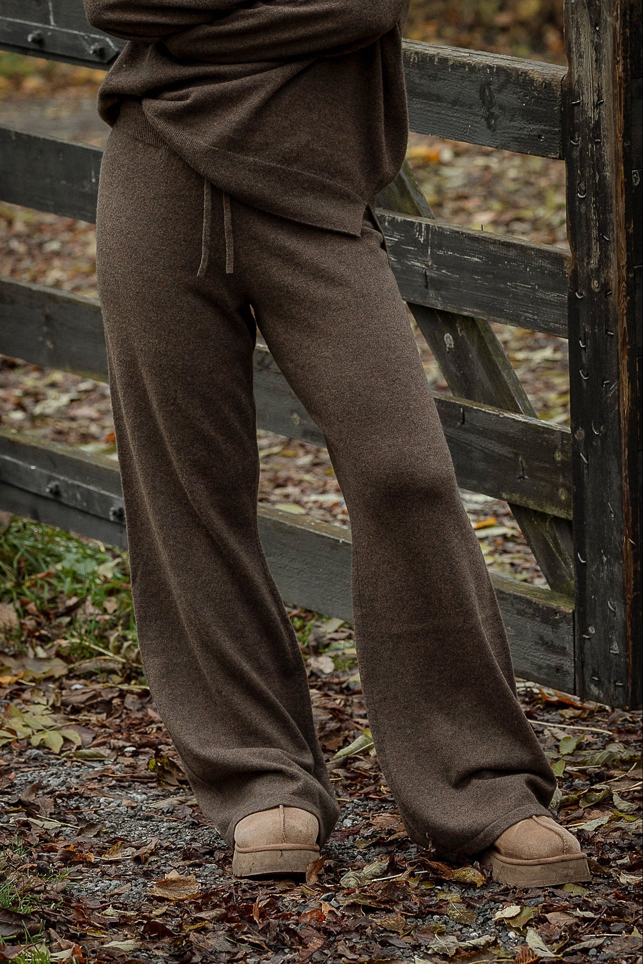 Person wearing brown pants standing next to a wooden fence in an outdoor setting with fallen leaves on the ground.
