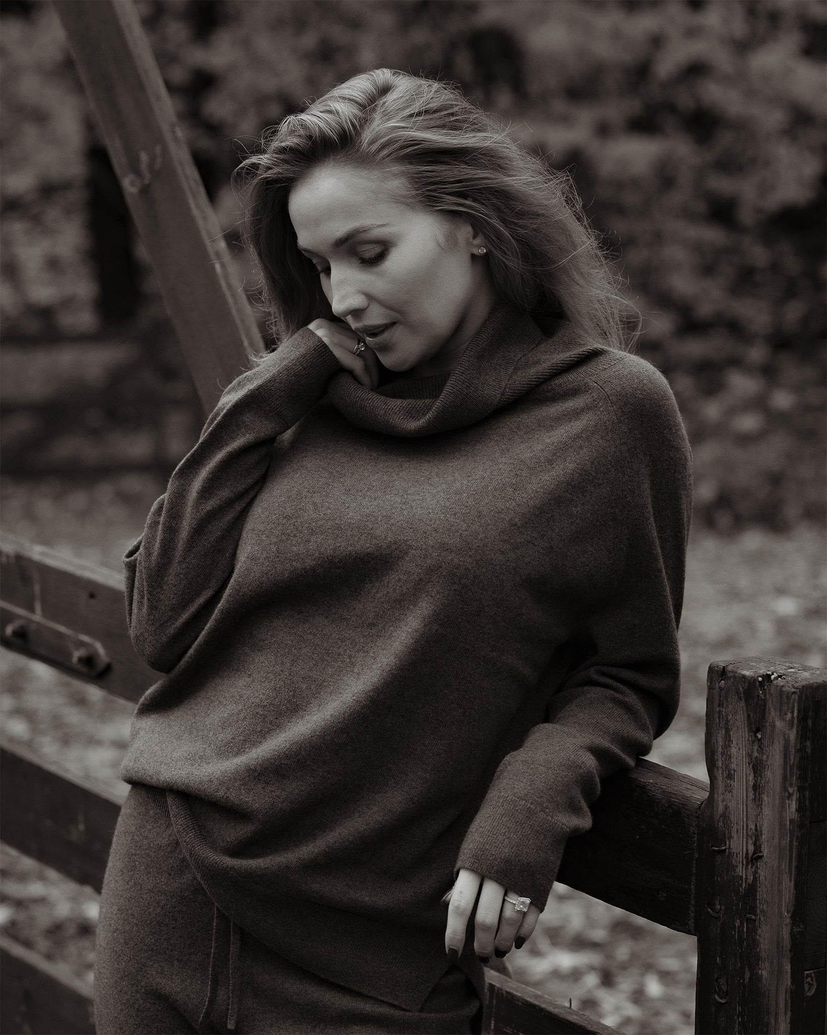Woman wearing a brown sweater leaning against a wooden fence in a natural setting