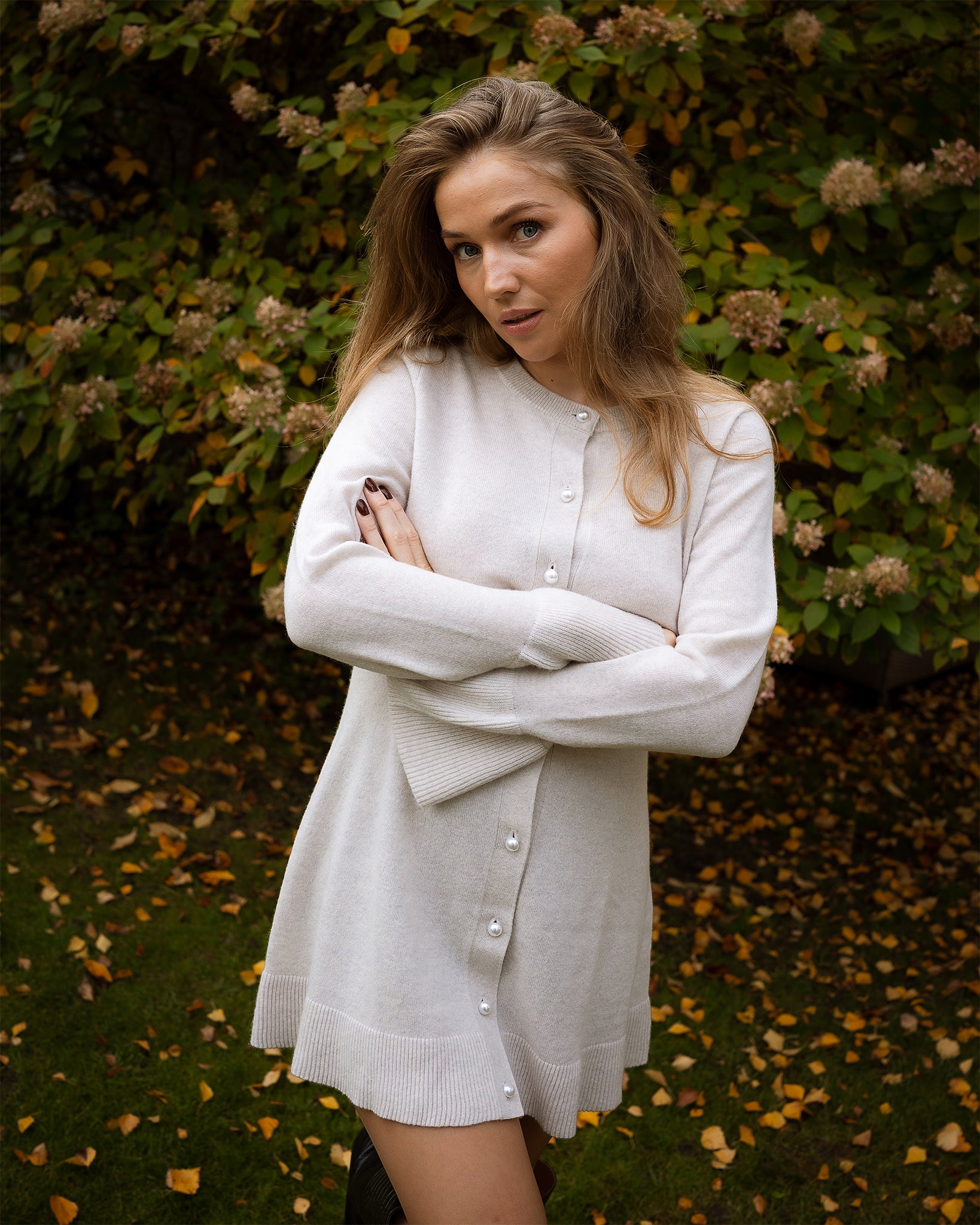 Woman wearing a white cardigan standing in front of green foliage with fallen leaves on the ground.