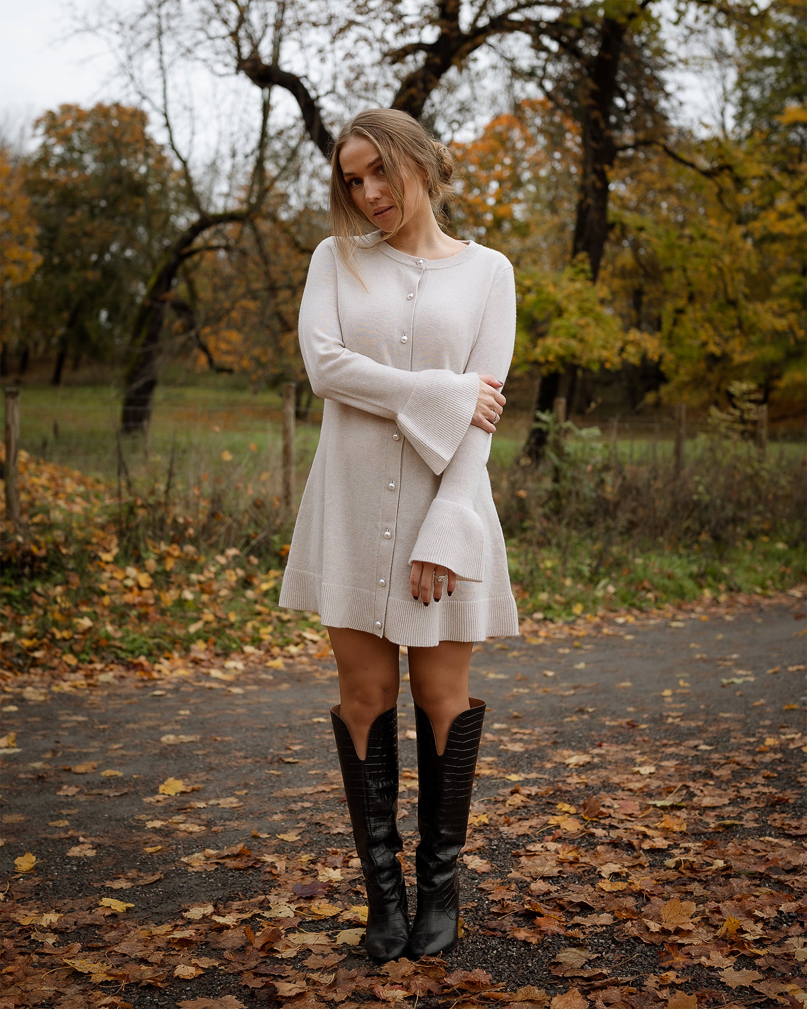 Woman in a white dress and black boots standing on a path with autumn leaves and trees.