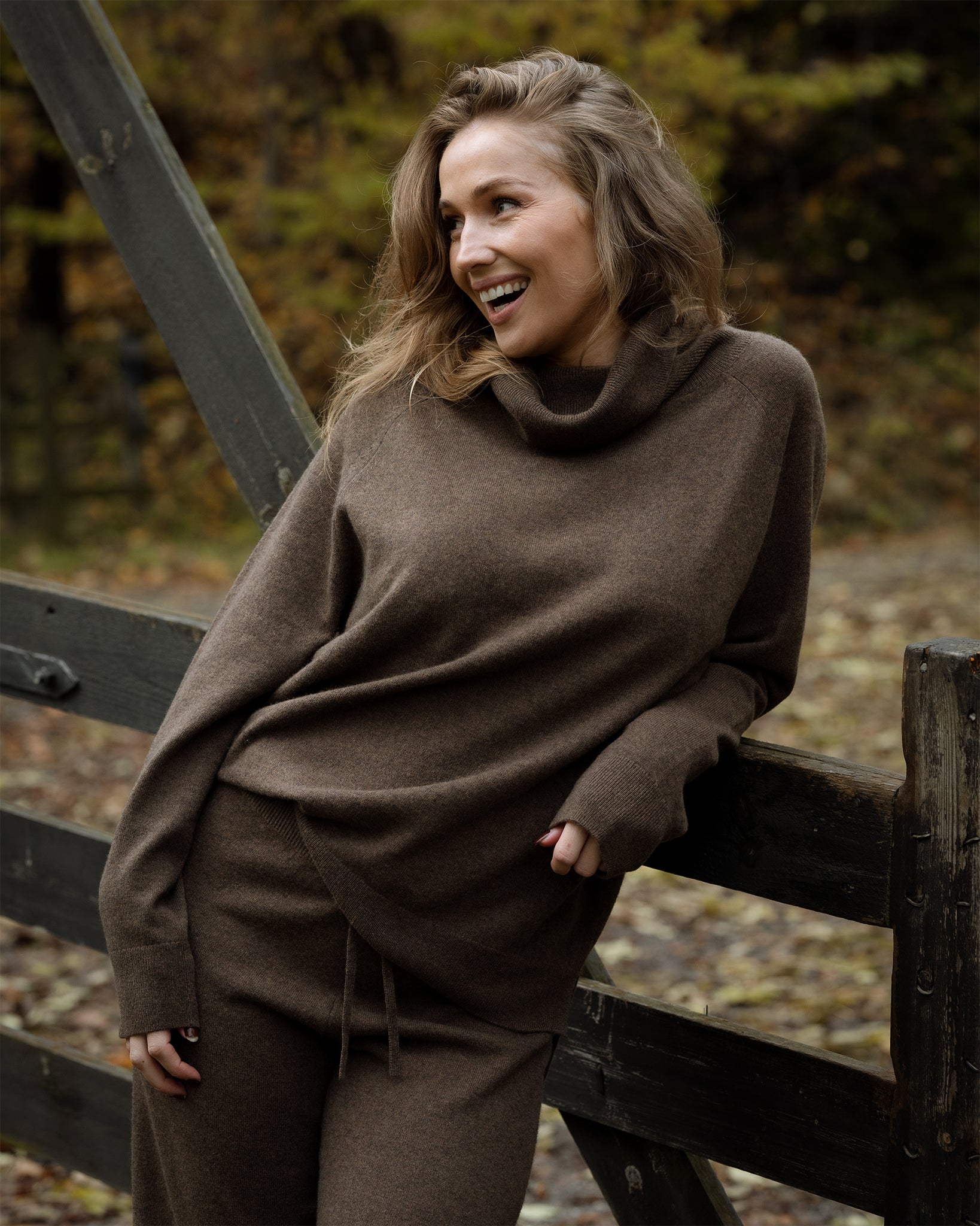 Woman wearing a brown sweater and pants leaning against a wooden fence in a forest setting.