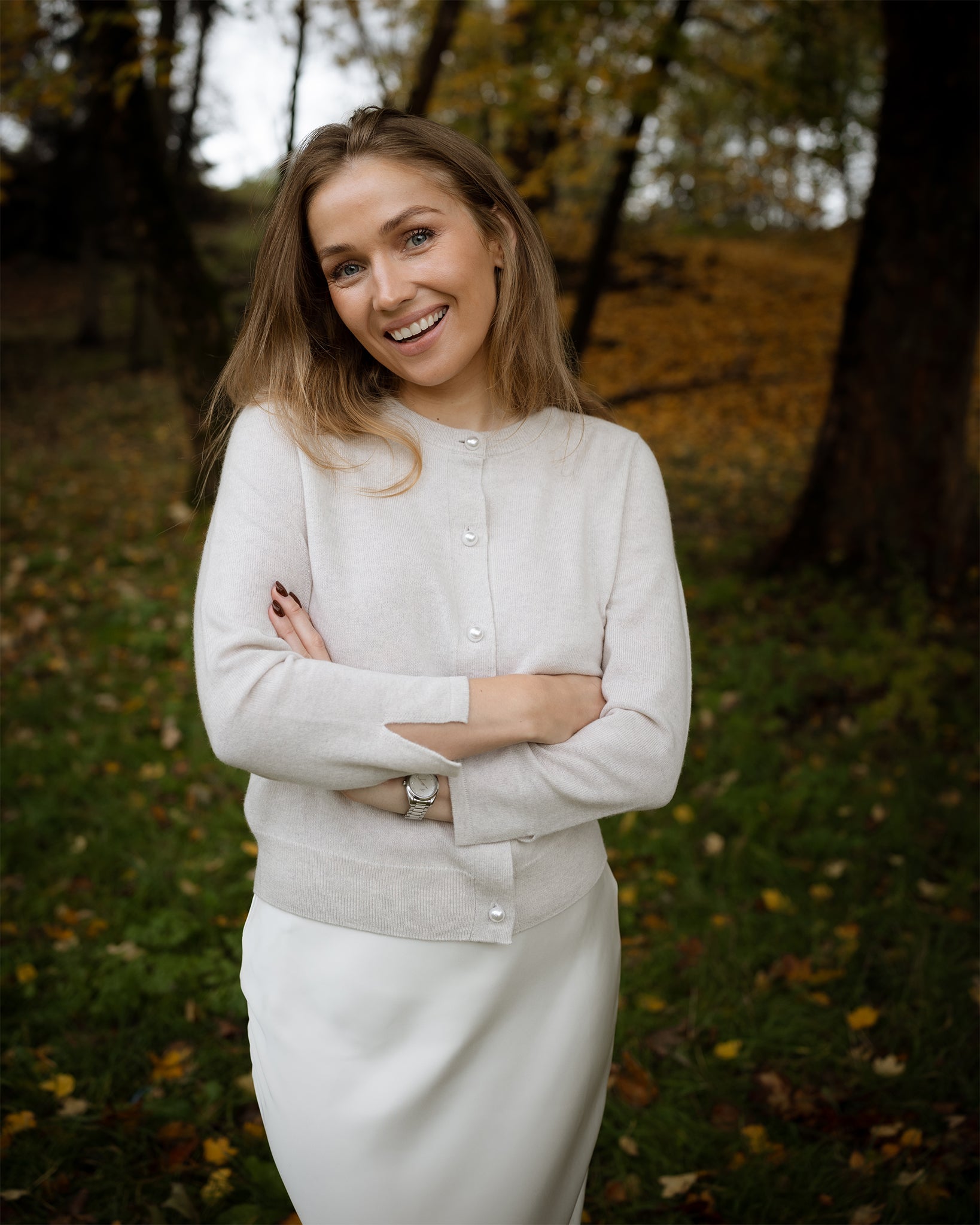 Woman in a light-colored outfit standing in a natural setting with autumn trees and grass.