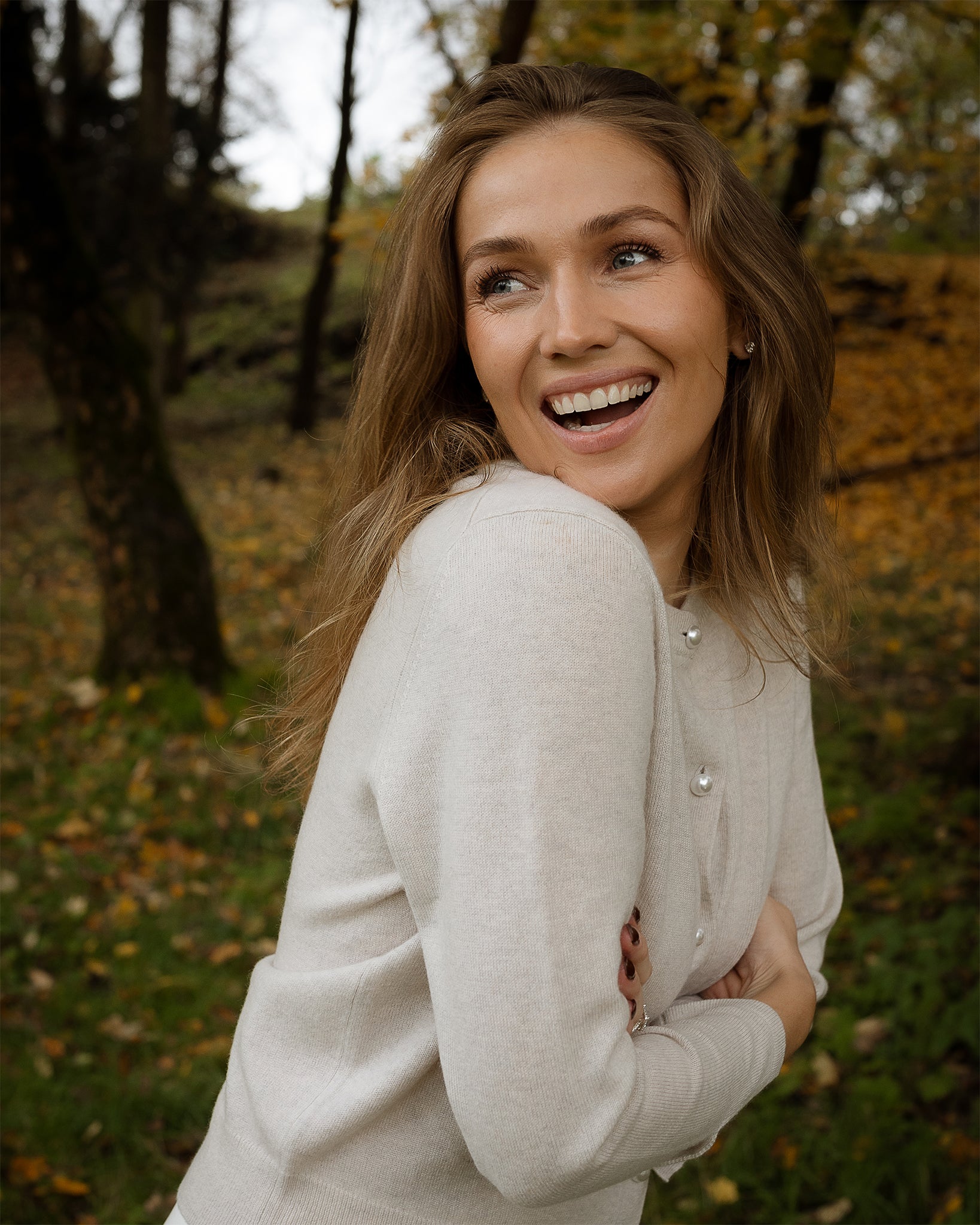 Woman in a beige sweater standing in an outdoor setting with trees and grass.