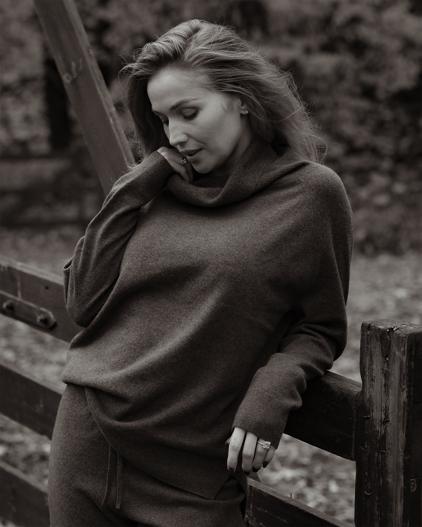 Woman wearing a brown sweater leaning against a wooden fence outdoors.